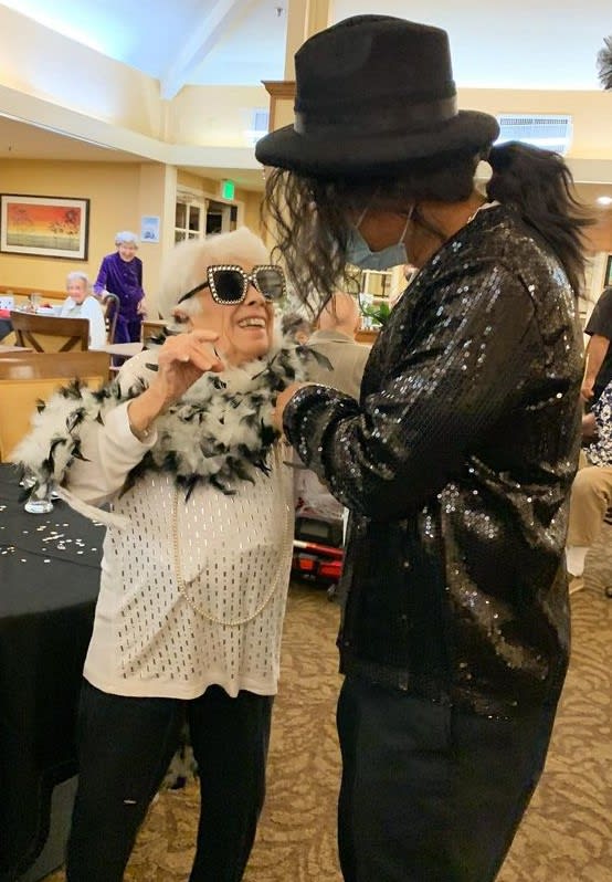 An elderly woman in stylish sunglasses and a feather boa dances joyfully with a partner at a festive gathering.