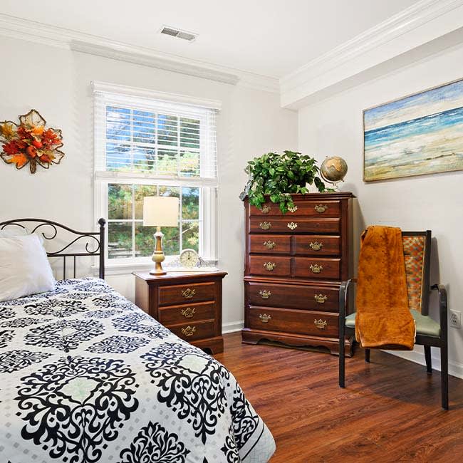A cozy bedroom with a metal frame bed, decorative bedding, and wooden dresser adorned with a plant and globe. A window lets in natural light, while artwork and an orange throw add warmth to the space.