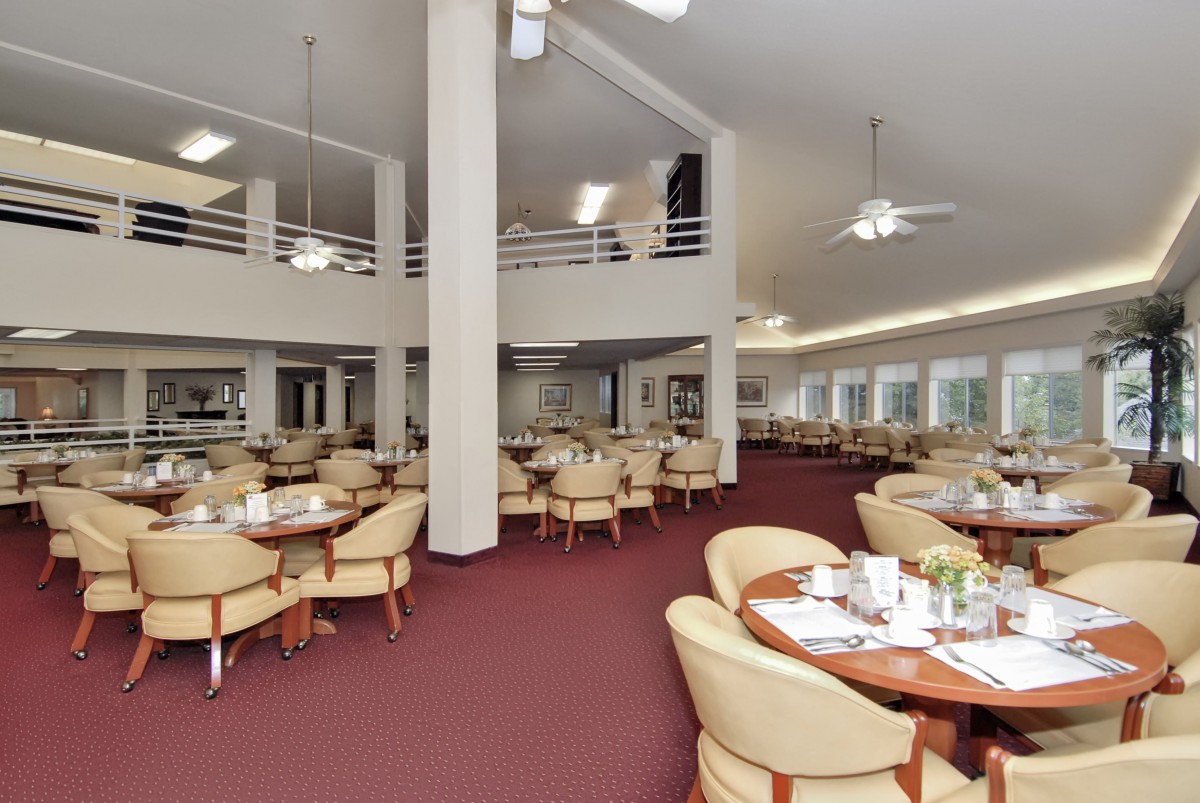 A spacious dining area with elegantly arranged tables and chairs, featuring a carpeted floor and ceiling fans.