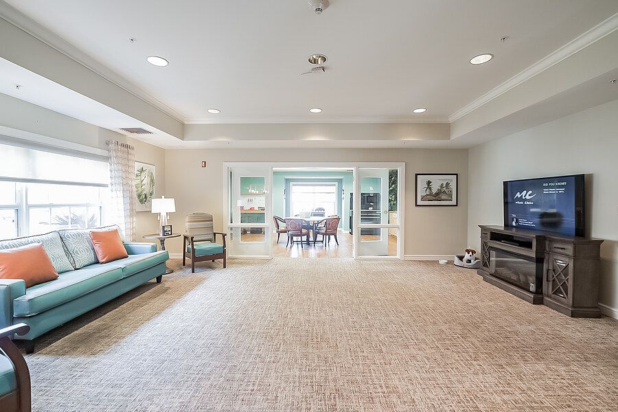 A spacious living room with a carpeted floor, featuring comfortable seating and a television, leading into a dining area.