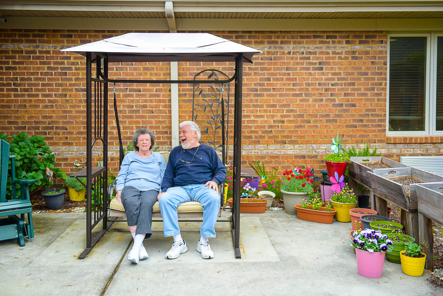 A couple sitting on a swing surrounded by colorful potted flowers in a garden area.