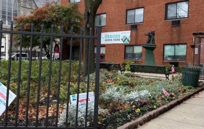 A community garden located in an urban setting, featuring a variety of plants and flowers, bordered by a metal fence and adjacent to a brick building.