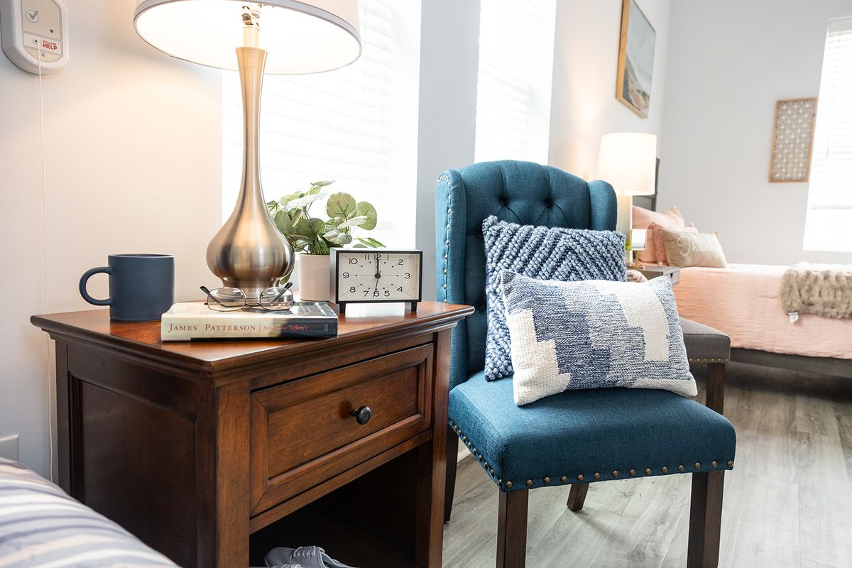 A cozy living room corner featuring a blue upholstered chair with decorative pillows, next to a wooden side table adorned with a lamp and small clock.