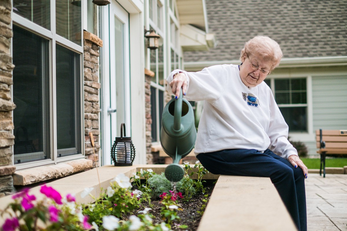 An elderly woman waters flowers in her garden on a sunny day.