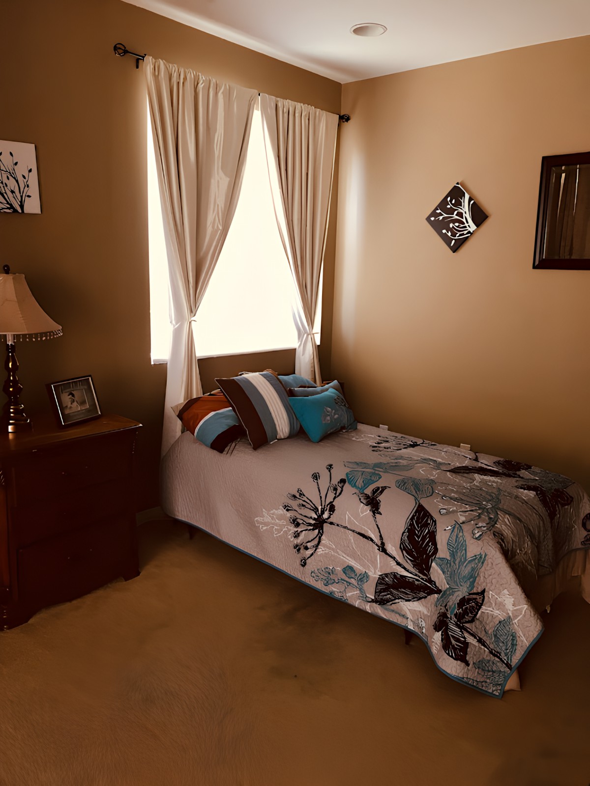 A cozy bedroom with a single bed adorned with decorative pillows and a floral comforter, beside a lamp on a nightstand, and framed wall art.