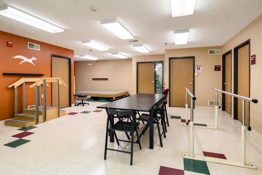 A rehabilitation facility room with therapy equipment, tables, and chairs arranged under bright fluorescent lighting.
