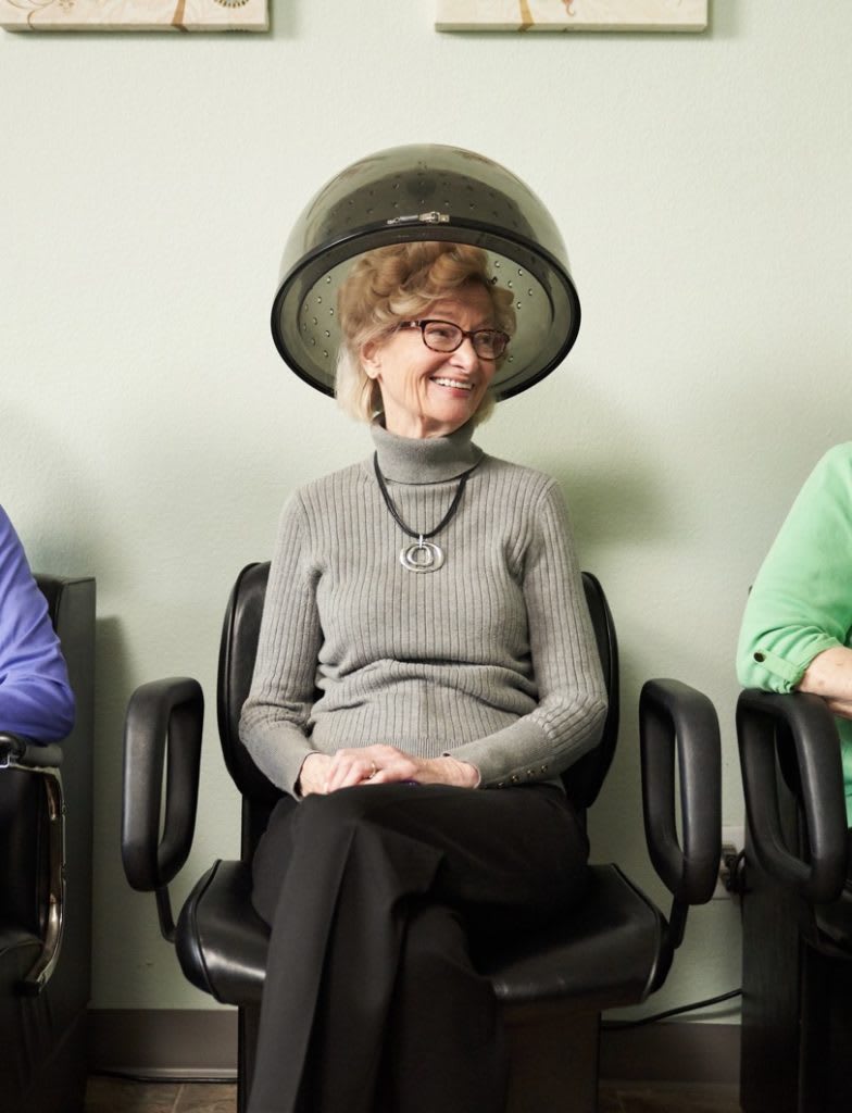 A woman sitting under a hair dryer at a salon, smiling and relaxed.
