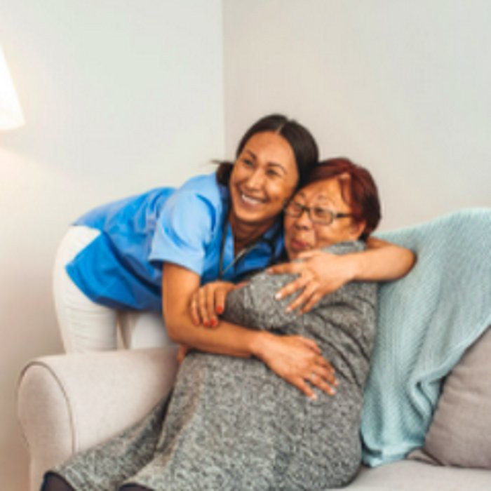 A caregiver warmly embraces an elderly woman on the couch, highlighting a moment of affection and support.