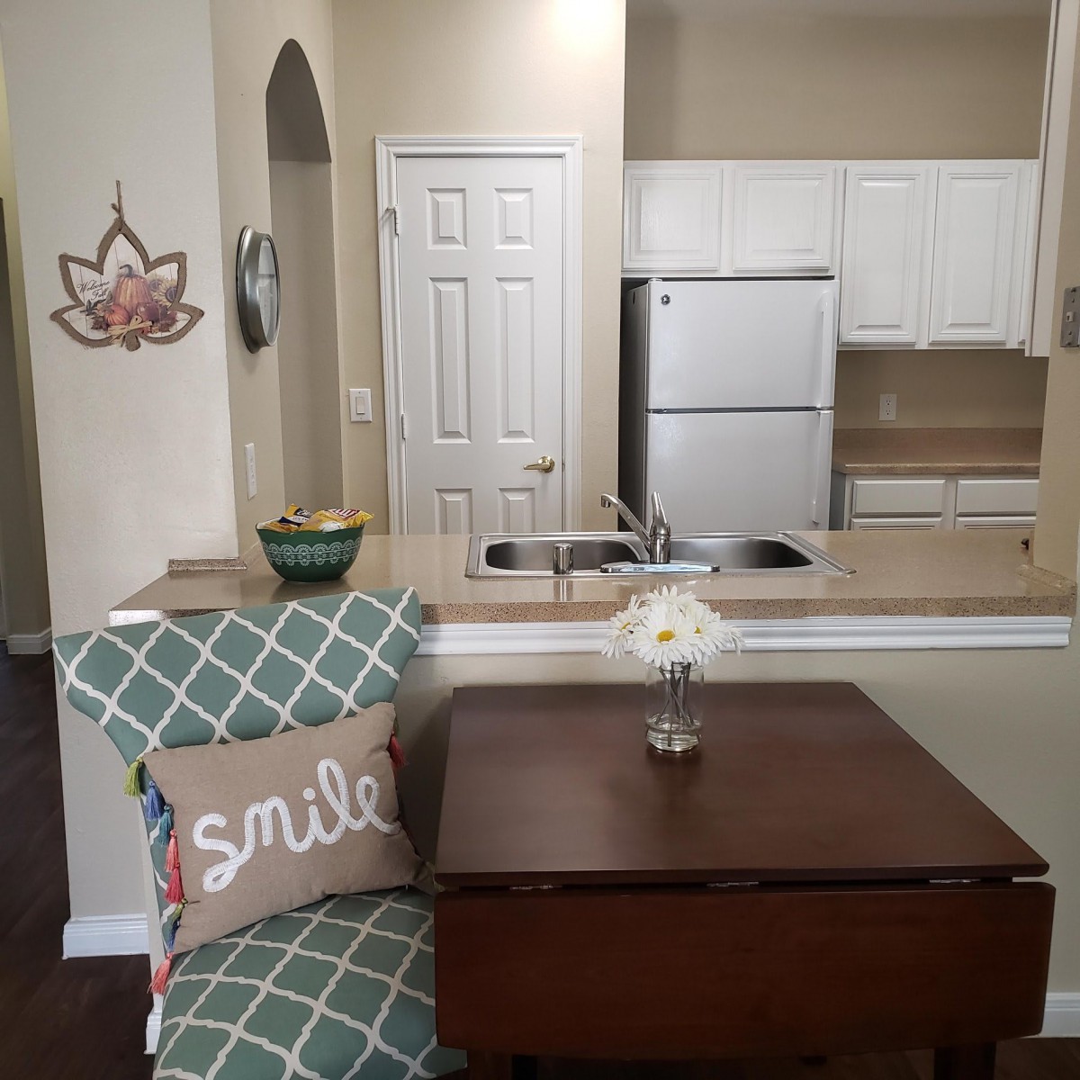 A cozy kitchen and dining area with modern touches, featuring a chair with a 'smile' pillow, a small table, and a flower vase.