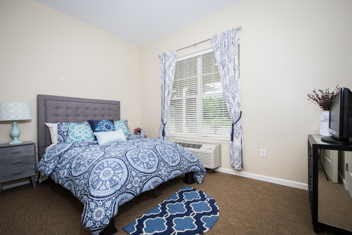 A cozy bedroom featuring blue patterned bedding and curtains, with a window allowing natural light to enter.