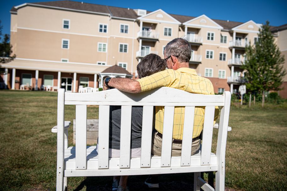 An elderly couple sits closely on a bench outside a retirement home.