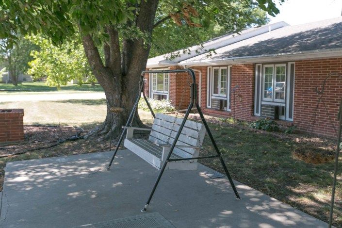 A swing bench under a large tree on a shaded patio in front of a house.