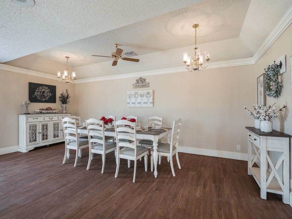 A spacious dining room with a large table and chairs, adorned with chandeliers and a ceiling fan, featuring wood flooring and decorative elements.
