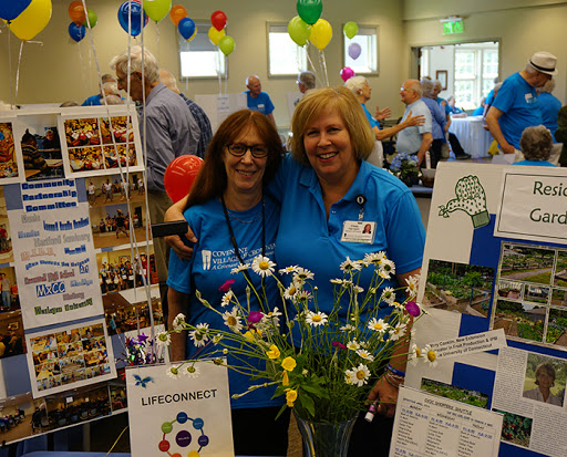 Two volunteers at a community event, smiling by their booth decorated with flowers and balloons.