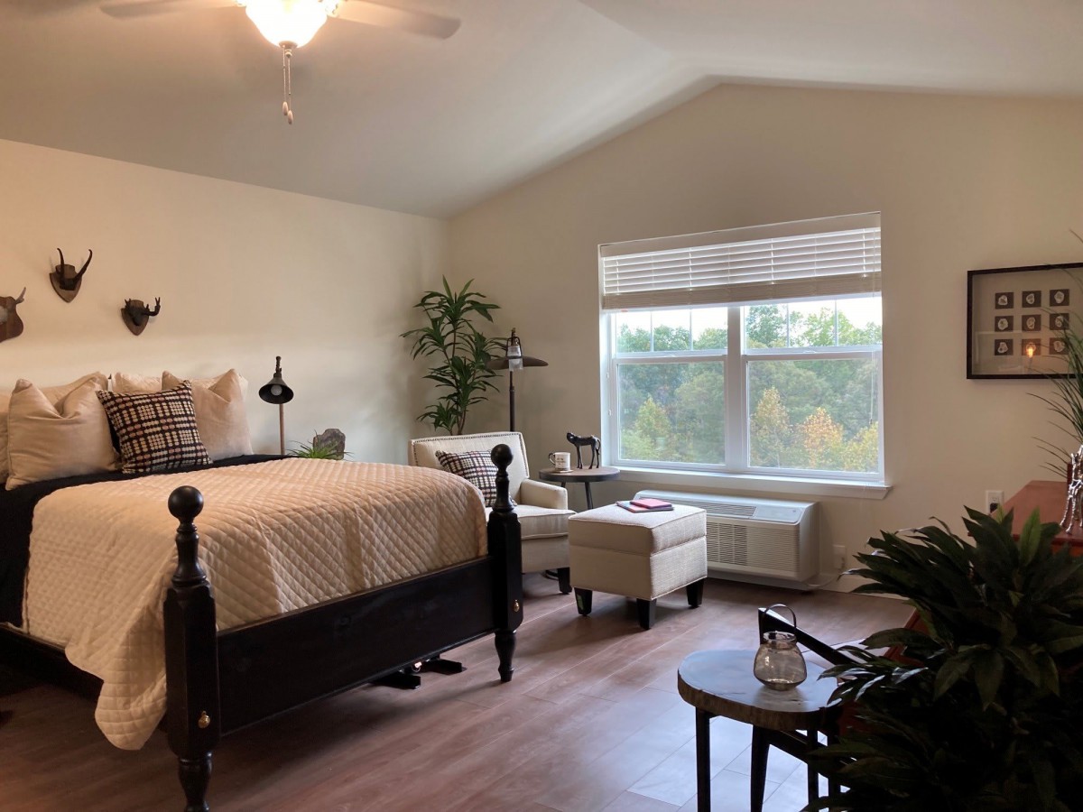 A cozy bedroom interior with a large bed, chairs by the window, and indoor plants.