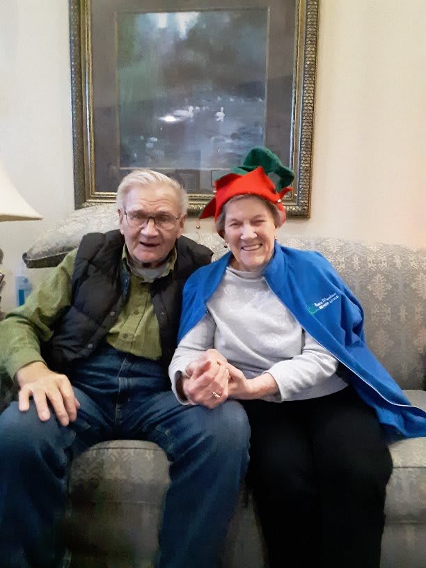 An elderly couple smiling and sitting together indoors, with the woman wearing a festive hat.