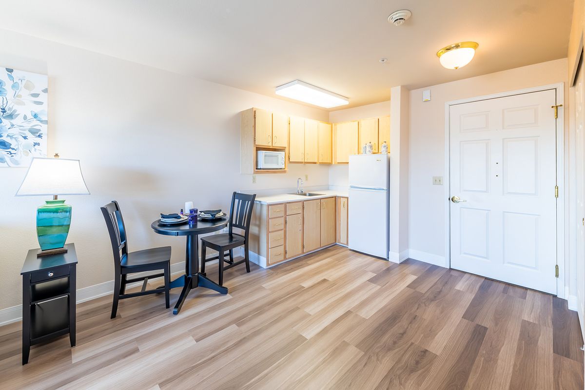 A compact kitchen and dining area featuring wooden cabinetry, a small round table with chairs, and bright natural lighting.