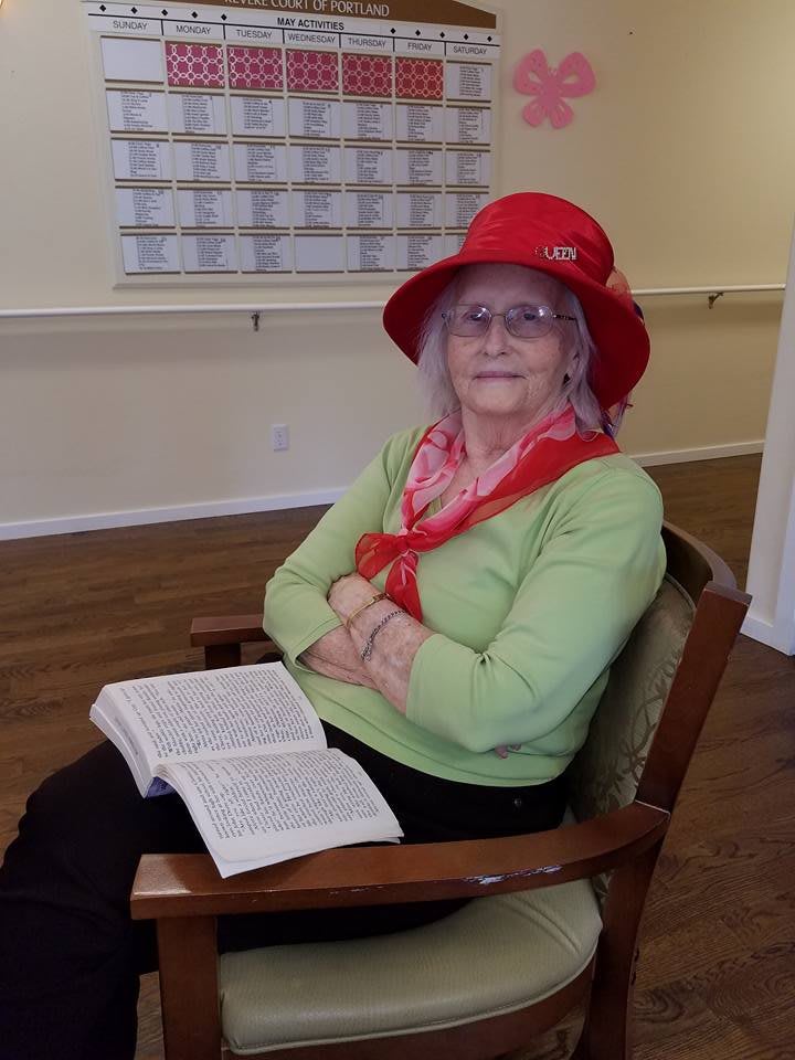 An elderly woman in a red hat and green sweater sitting on a chair with an open book.