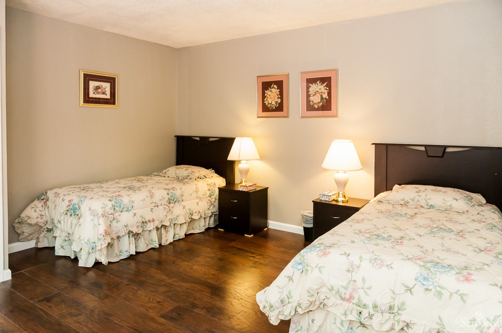 A cozy bedroom with two single beds, nightstands, and lamps, featuring hardwood flooring and framed artwork on the walls.