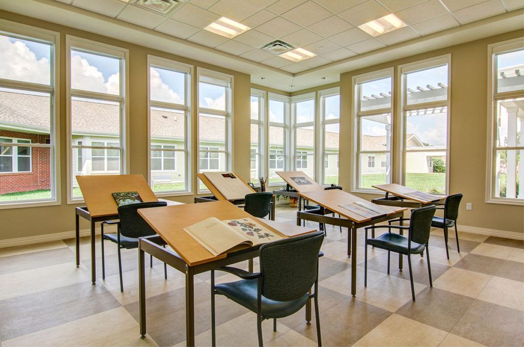 A bright classroom with art desks and chairs arranged neatly by large windows.