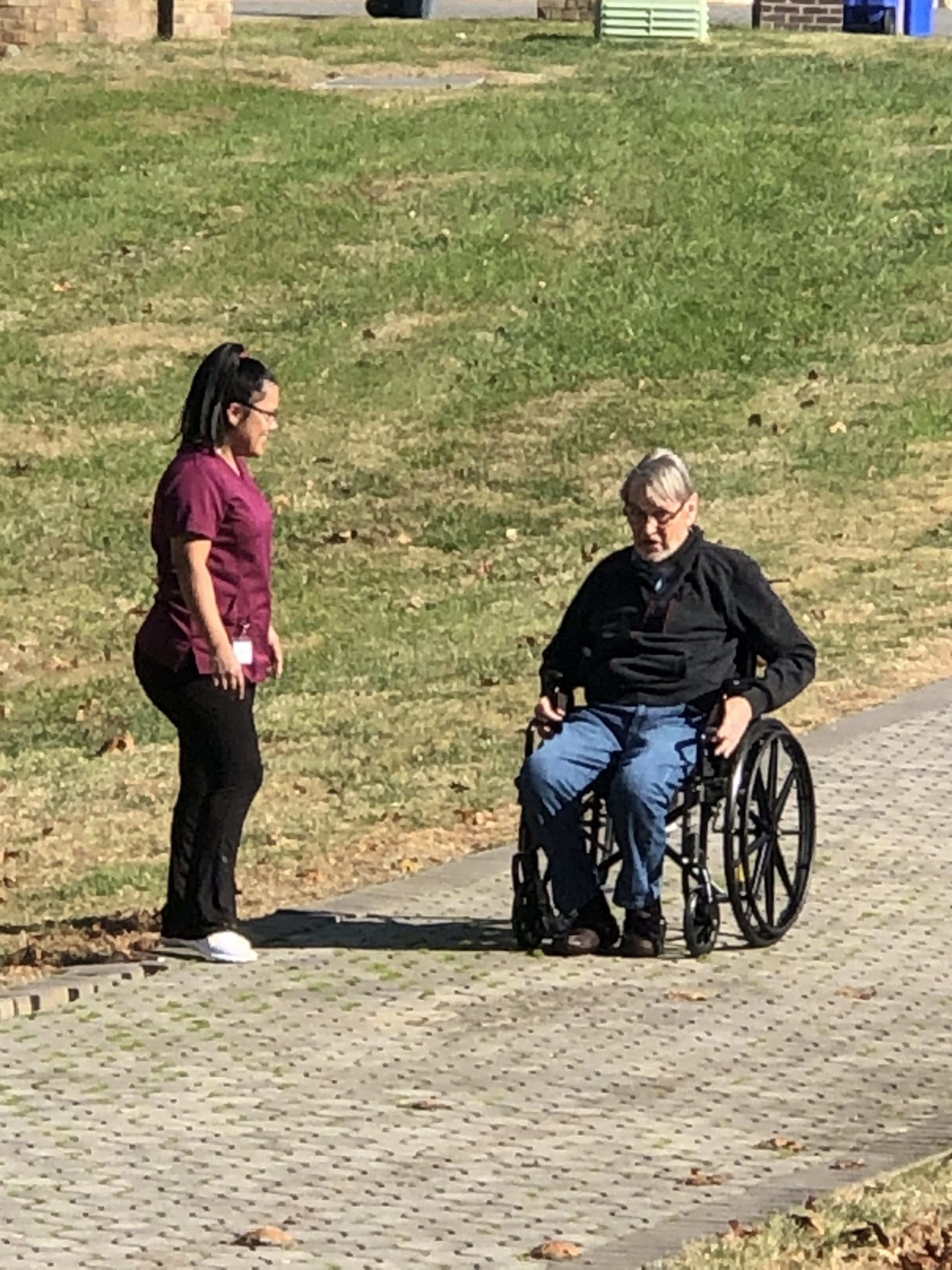 A caretaker interacts with an elderly person in a wheelchair on a sunny day.