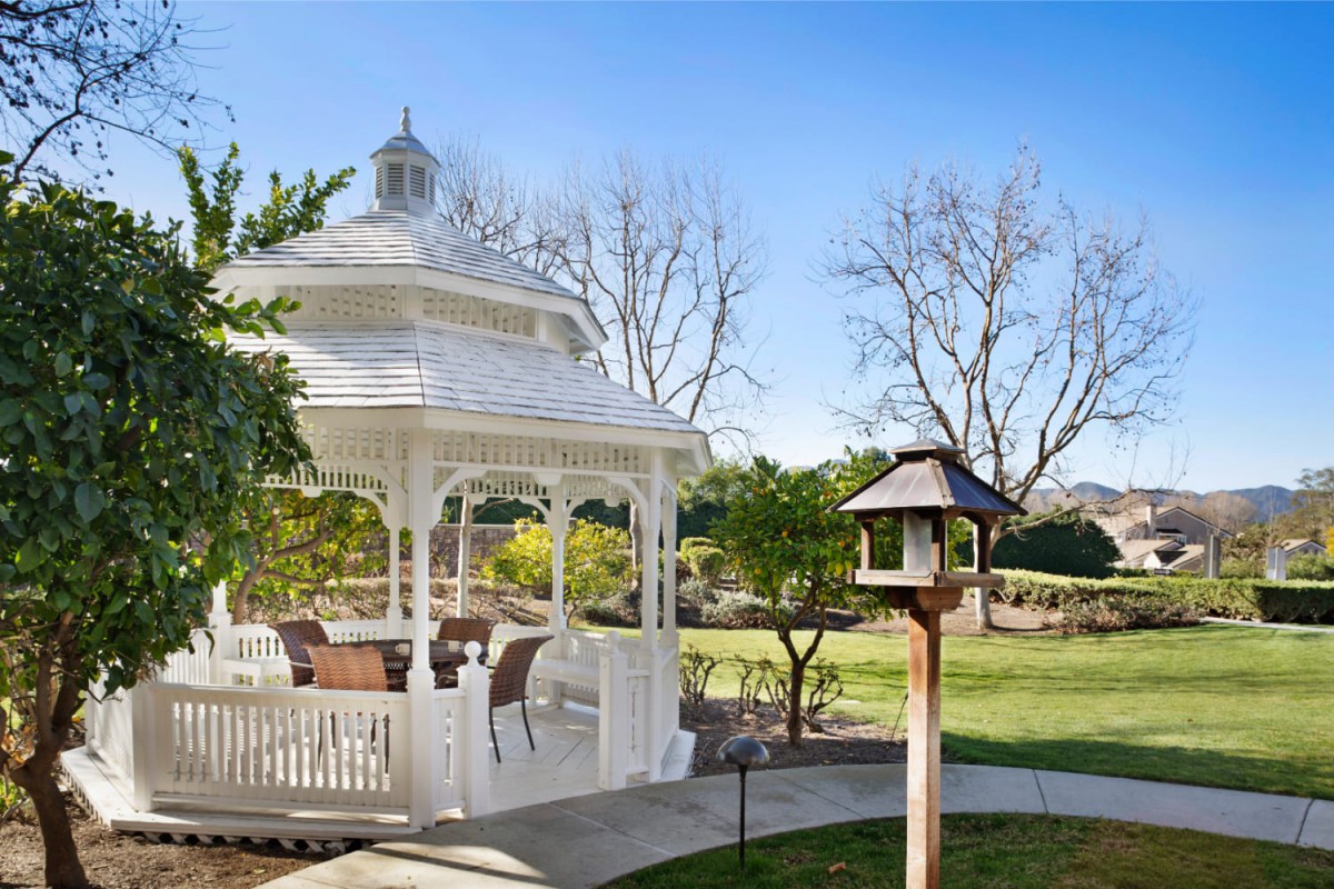 A picturesque garden scene featuring a white gazebo surrounded by greenery and a birdhouse on a sunny day.