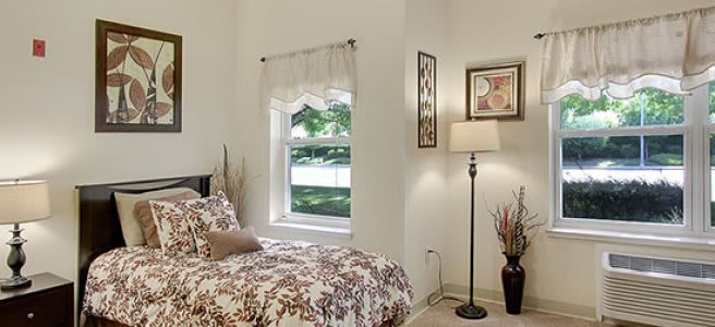 A cozy bedroom with a floral bedspread, standing lamp, wall art, and natural light streaming through the window.
