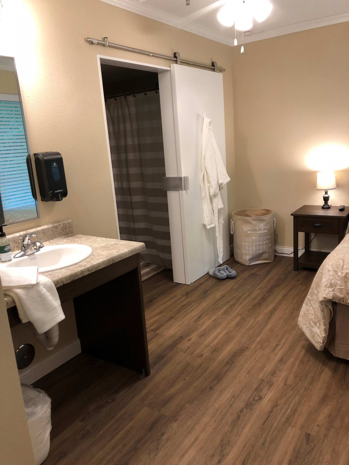 A cozy bedroom with an adjoining bathroom, featuring a bed, bedside table with a lamp, and a sink area.