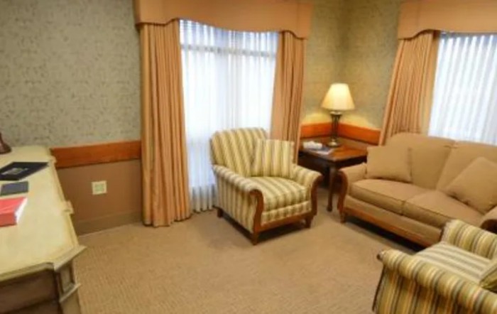 A cozy living room setup with a striped armchair, sofa, lamp on a side table, and draped curtains over a window.