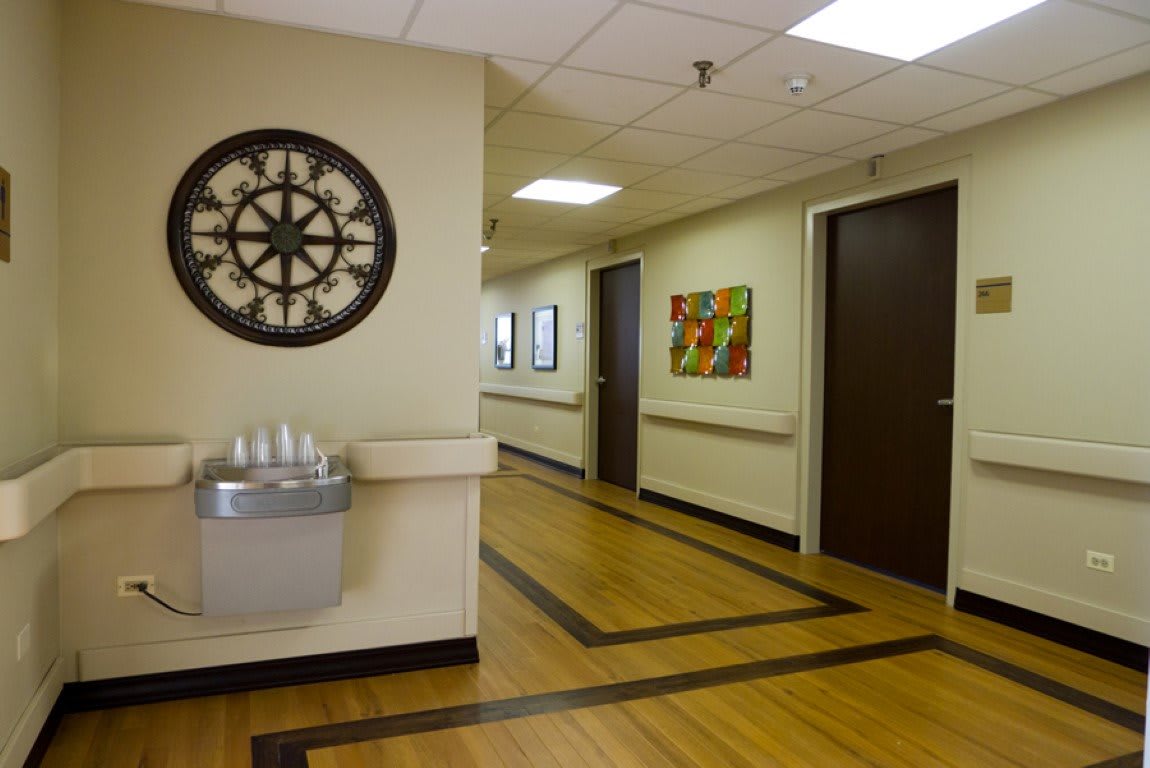 A hospital hallway with a clock, artwork on the wall, and a water fountain.