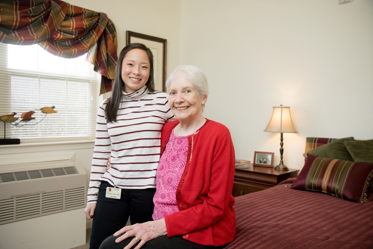 A young woman stands beside an elderly woman seated on a bed, both smiling warmly in a cozy bedroom setting.