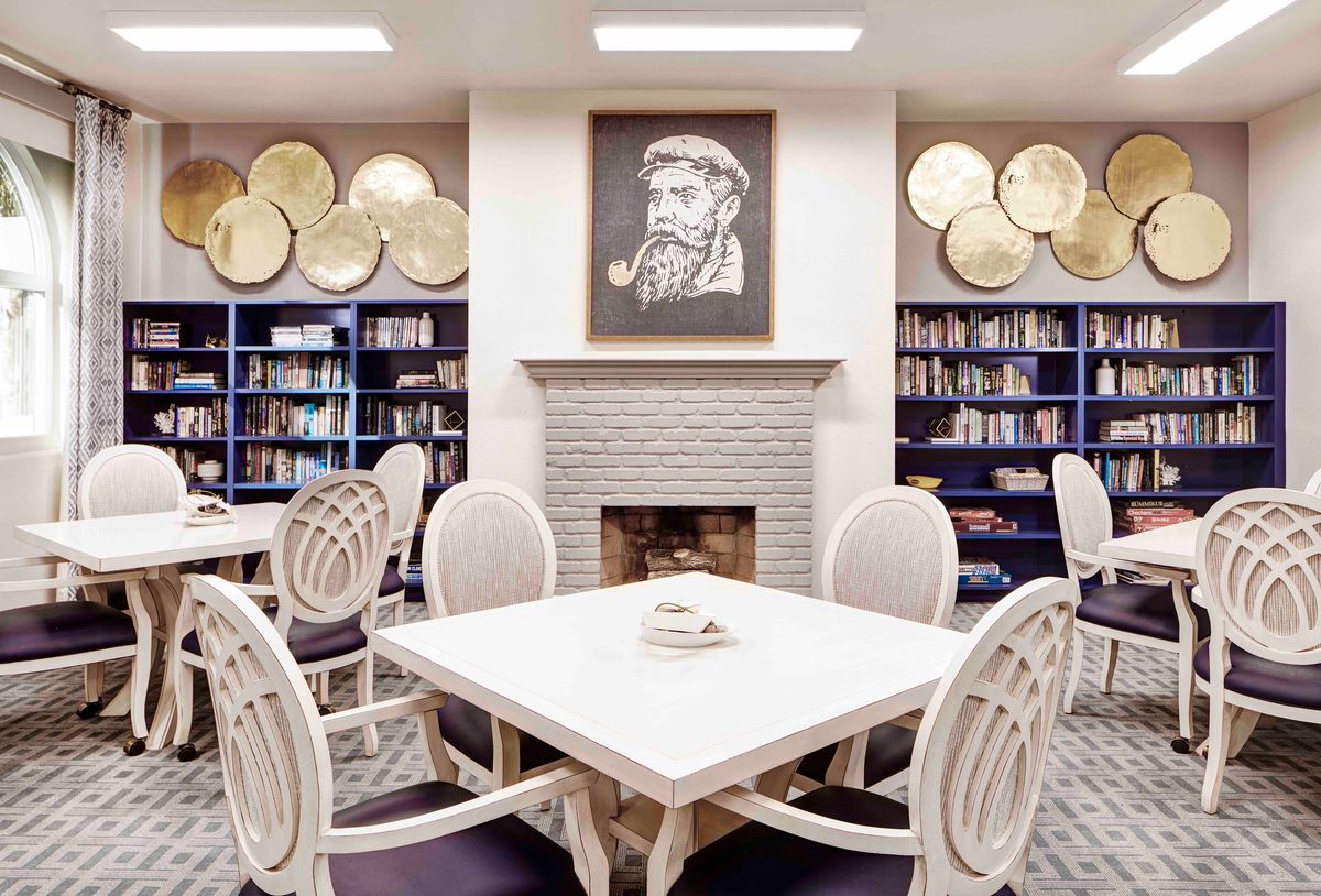 A cozy library with bookshelves, a central table with chairs, and a fireplace adorned with a portrait.