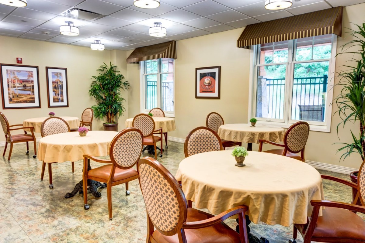 A cozy dining room with round tables, chairs, and indoor plants, well-lit with natural light from the windows.