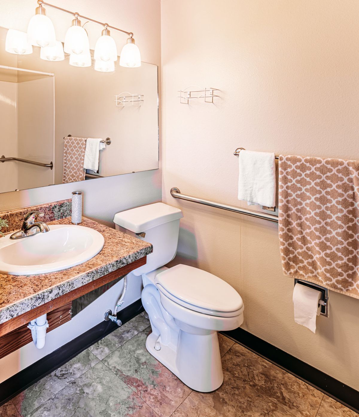 A clean and well-lit bathroom with granite countertops, featuring a sink, toilet, mirror, towel rack, and accessibility features.
