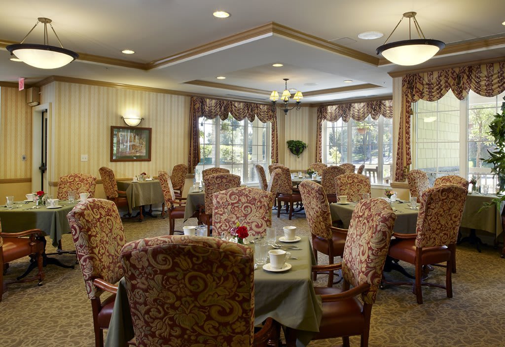 A cozy dining room with elegantly set tables and upholstered chairs, featuring warm lighting and classic decor.