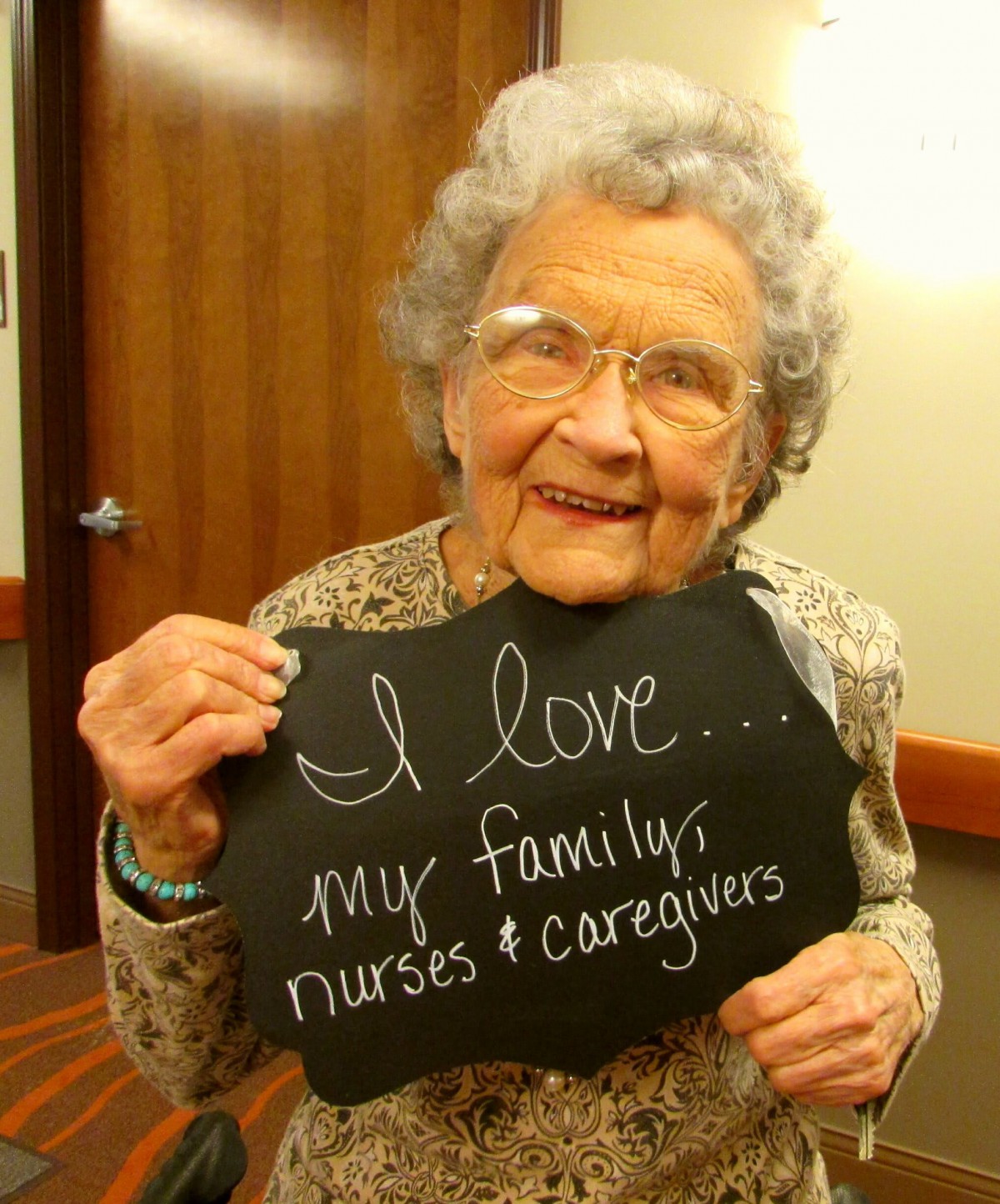 An elderly woman smiles while holding a sign expressing love for her family, nurses, and caregivers.