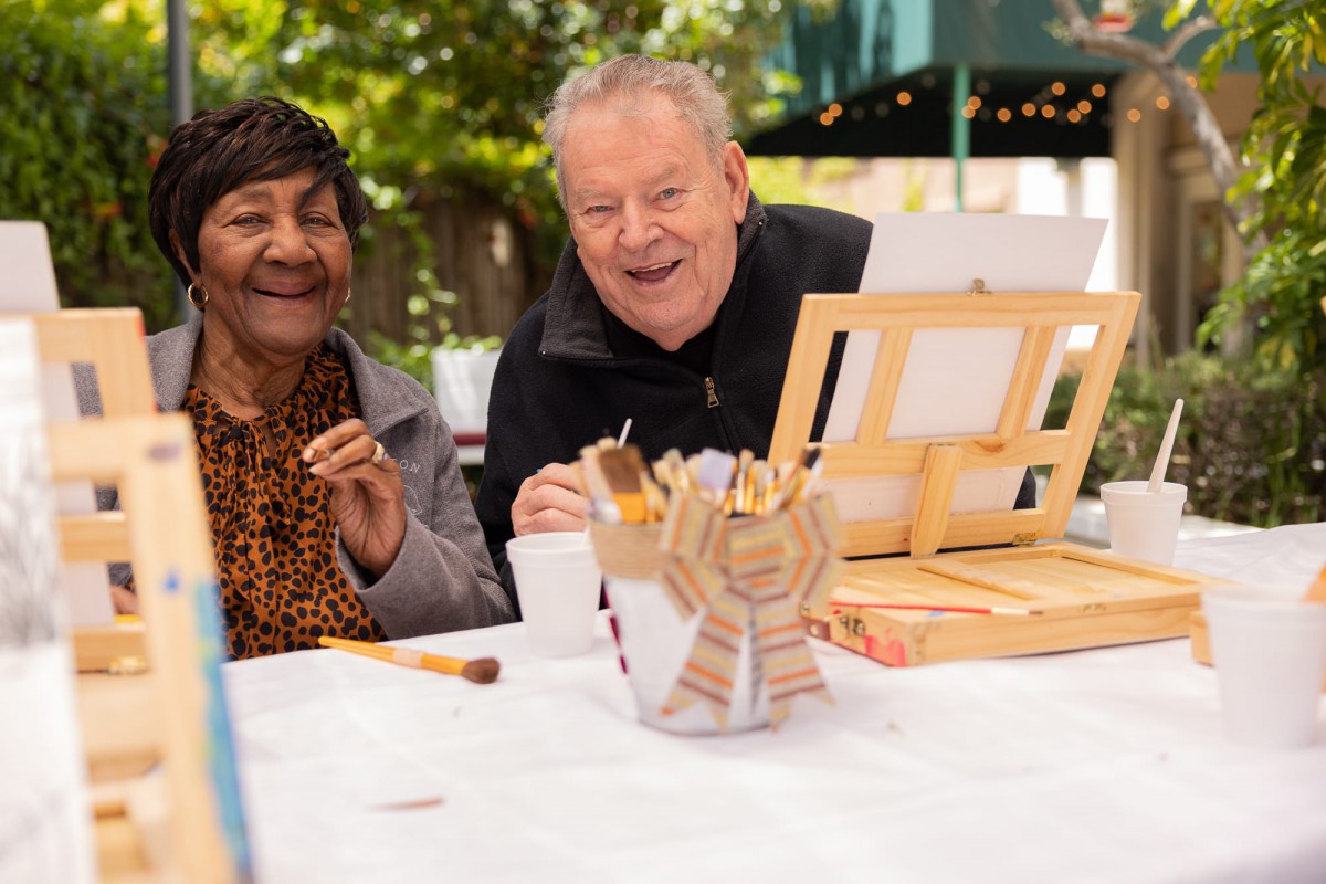 Two seniors enjoying an outdoor painting class together, smiling and engaged in their artwork.