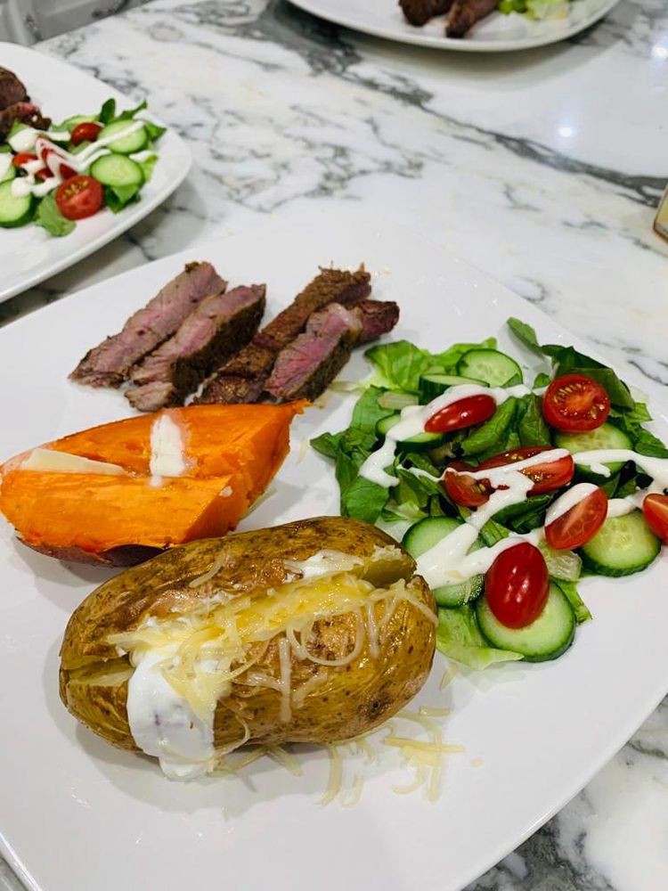A delicious meal featuring sliced steak, a baked potato topped with cheese and sour cream, sweet potato slices, and a fresh salad with cherry tomatoes and cucumber drizzled with ranch dressing.