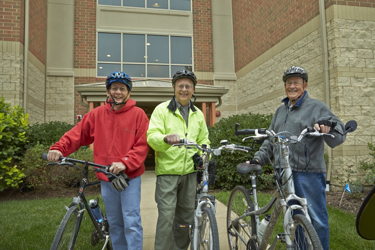 Three people with bicycles standing outside a building, wearing helmets and smiling.