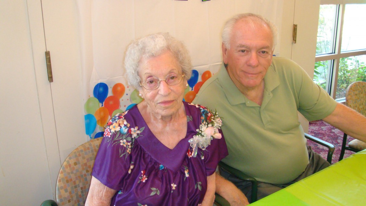 An elderly woman and a man sitting together at a celebration with colorful balloons in the background.