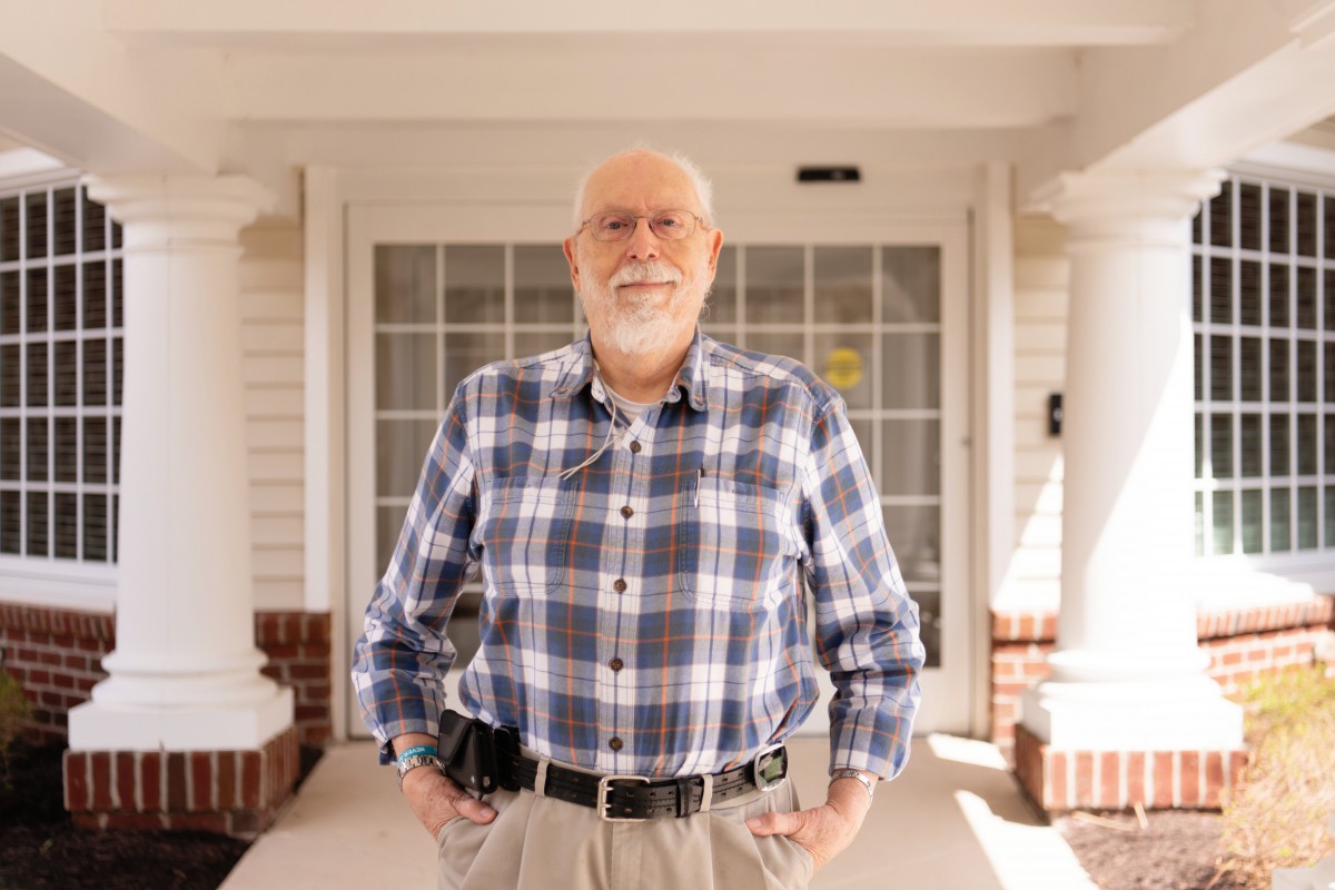 A smiling elderly man stands in front of a building, wearing a plaid shirt and glasses.