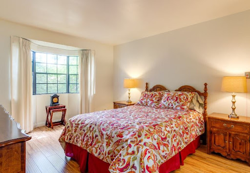 A cozy bedroom featuring a floral patterned bedspread, natural light from a large window, and warm wooden furniture.