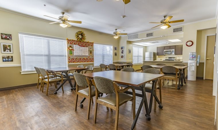 A well-lit communal dining area with multiple tables and chairs, ceiling fans, and large windows.