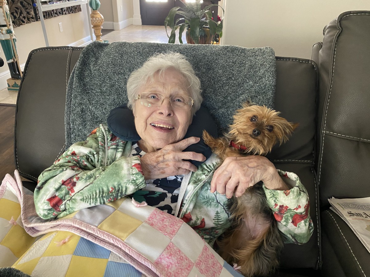 An elderly woman smiling while sitting on a couch with a small dog.