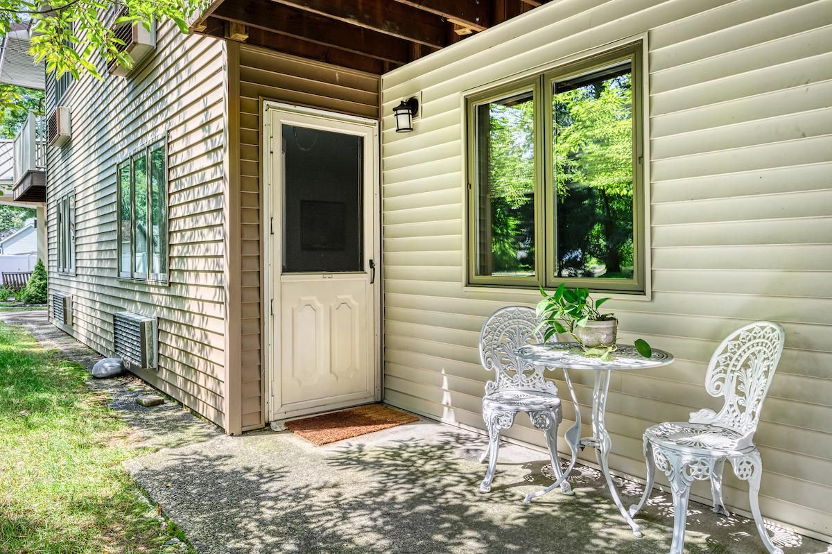 A cozy outdoor patio with a small table and chairs near a doorway, surrounded by greenery and dappled sunlight.