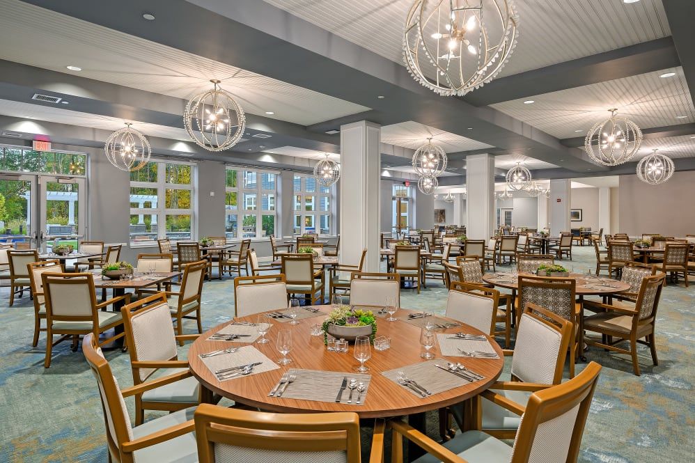 A spacious and elegantly designed dining area with round wooden tables, cushioned chairs, and modern chandeliers.