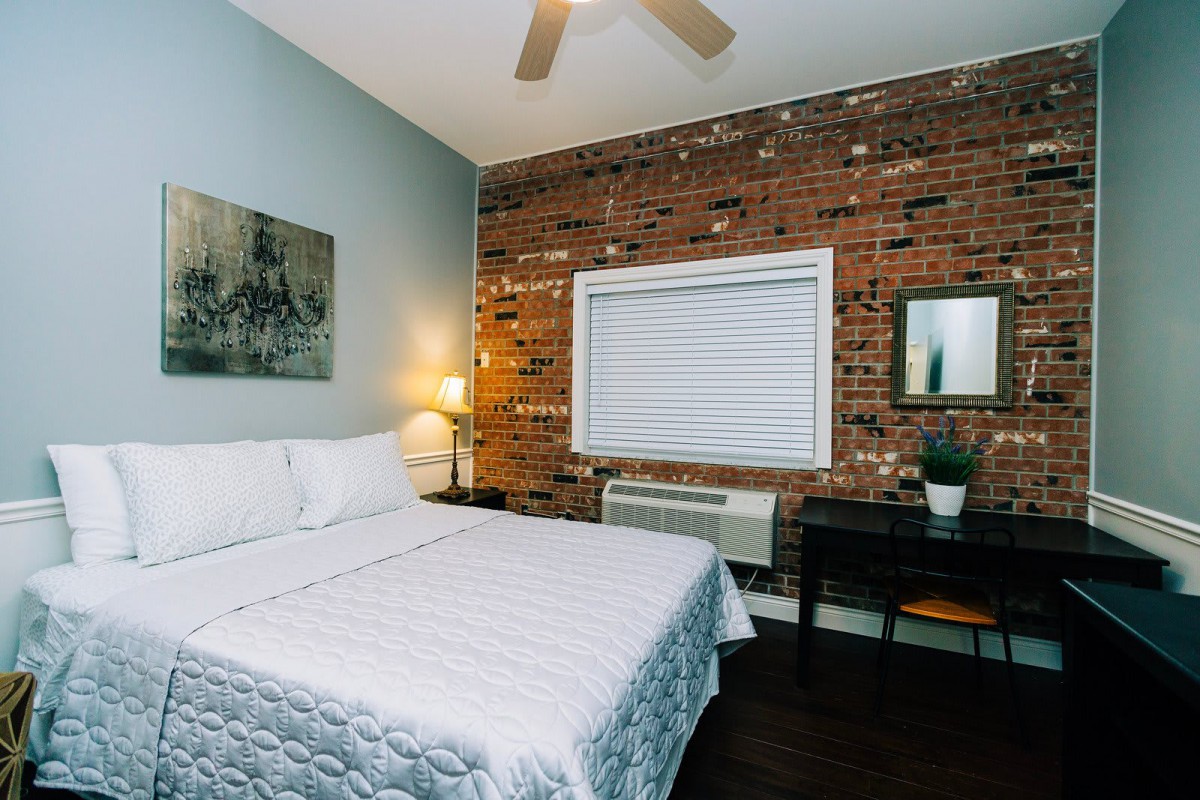 A cozy bedroom with a brick accent wall, featuring a bed with white linens, a lamp, and a small desk with a mirror.