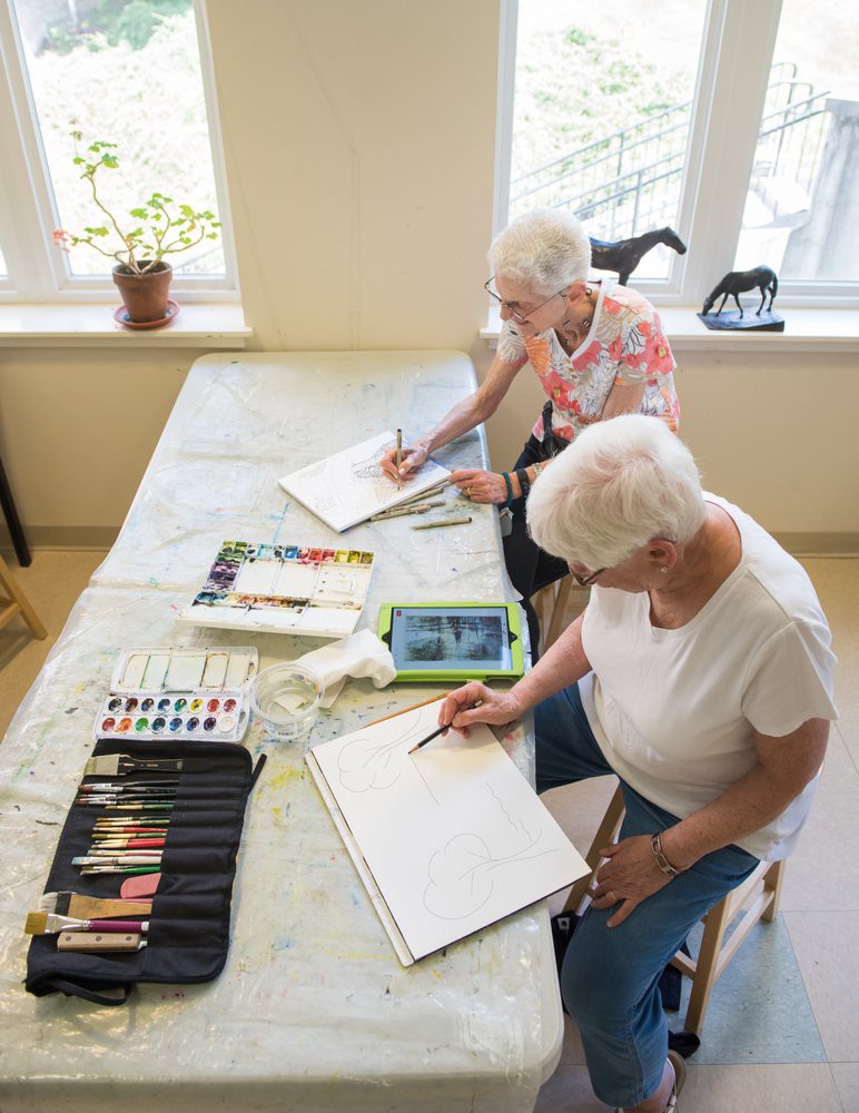 Two seniors engaged in a painting activity, surrounded by art supplies in a well-lit room.