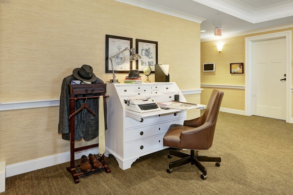 A vintage-style office setup featuring a white desk, brown chair, and a coat rack with a hat.