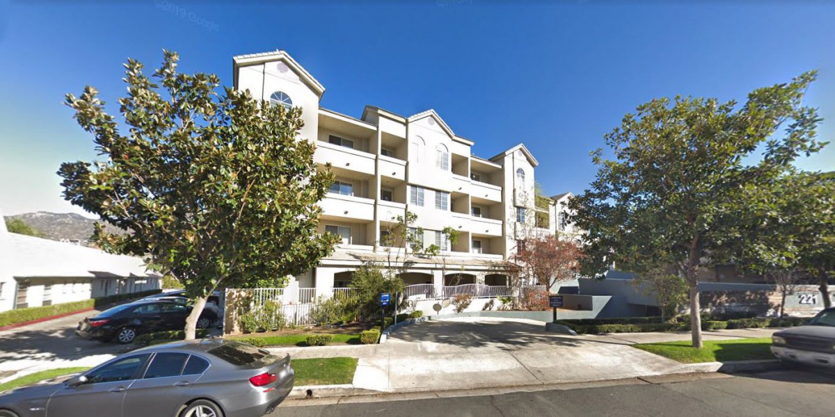 A modern apartment building with a driveway, surrounded by trees and parked cars on a sunny day.